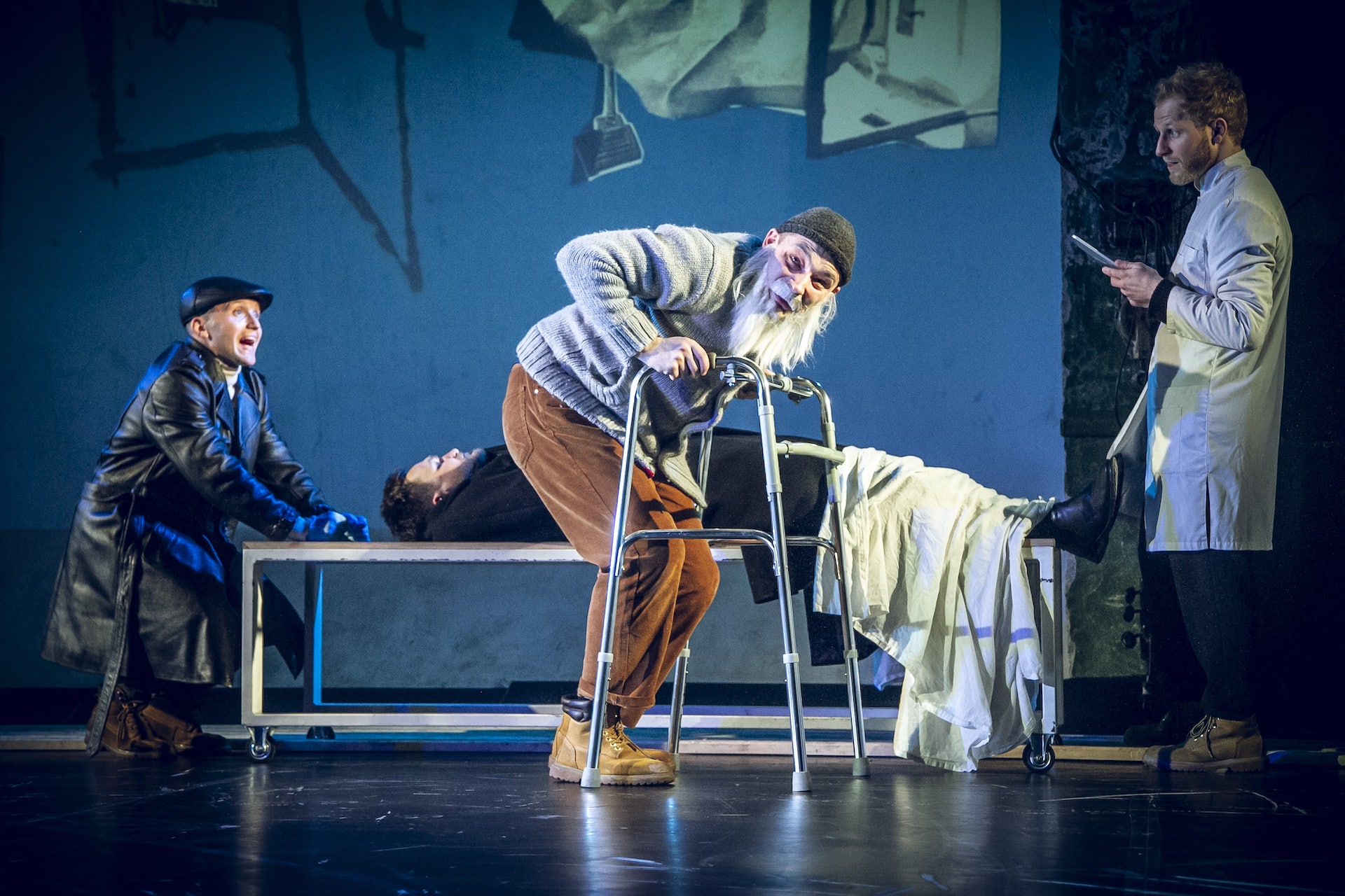 One performer dressed as an old man stands in the middle of the stage with a walker. One performer behind is lying on a bed, half covered with white blanket while the other two stands at each end of the bed.
