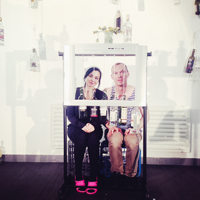 Two people sitting in front of a frame on which alcohol bottles are hanging