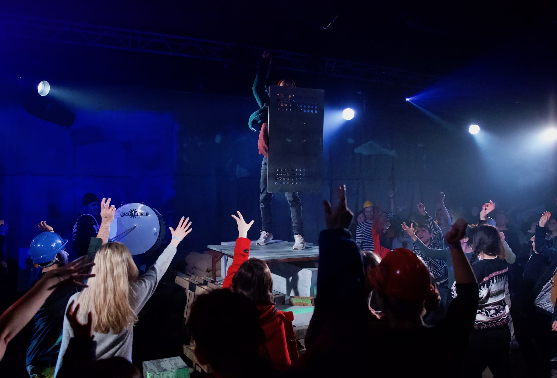 One performer standing in the middle holding a metal shield while the others stand around and holding their hands in the air