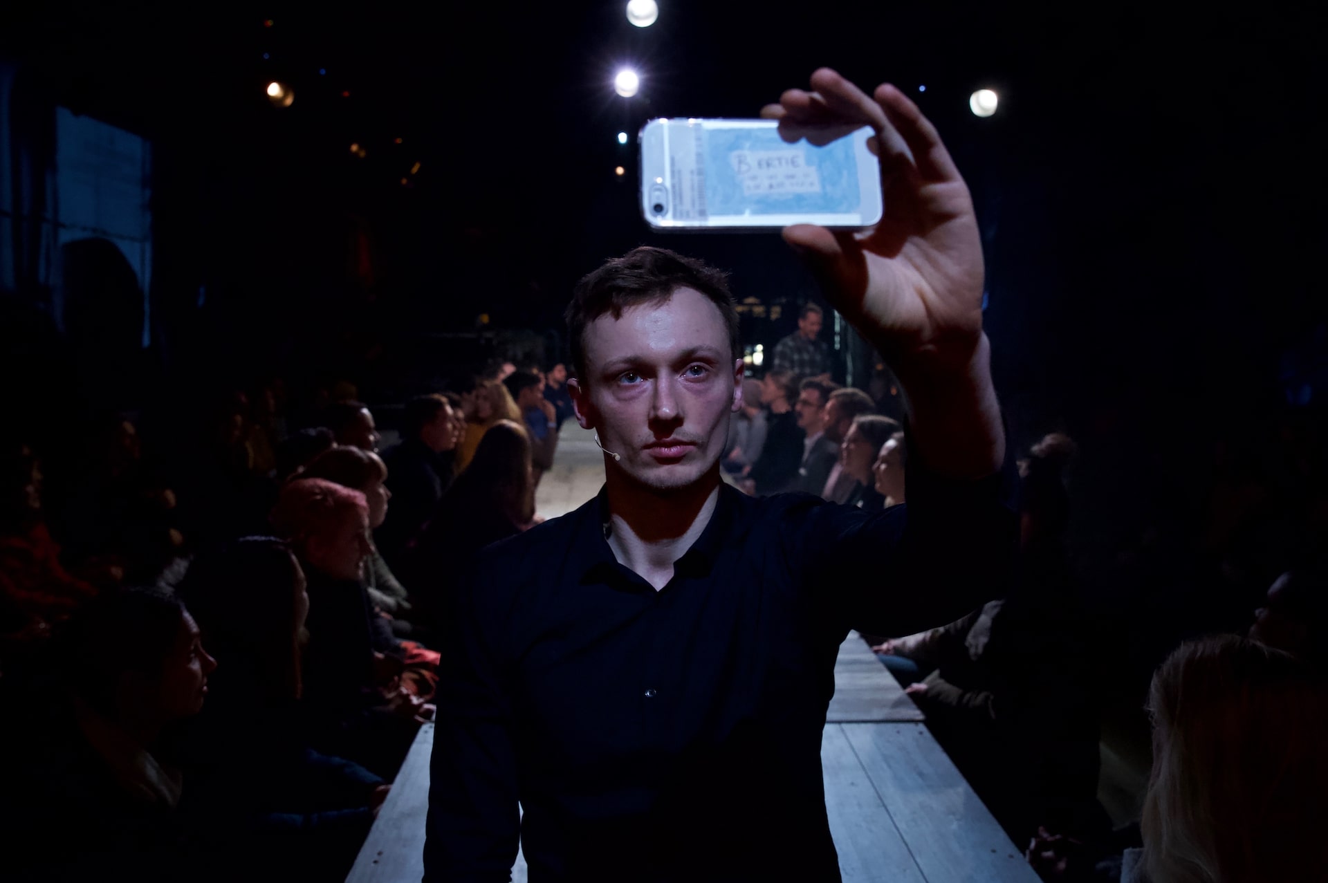 A performer standing on a long table, holding up a phone written bertie on the back