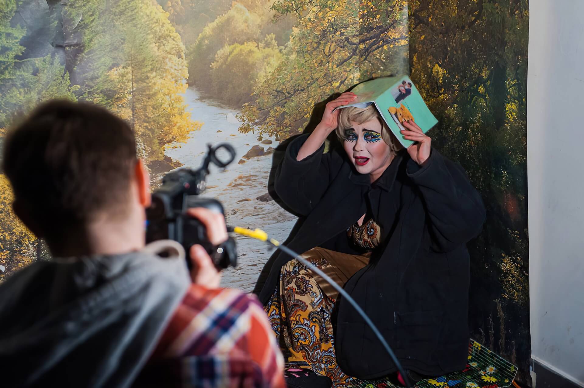 A person filming a woman with a heavy makeup. The woman looks scared and is holding up a book, covering her head.