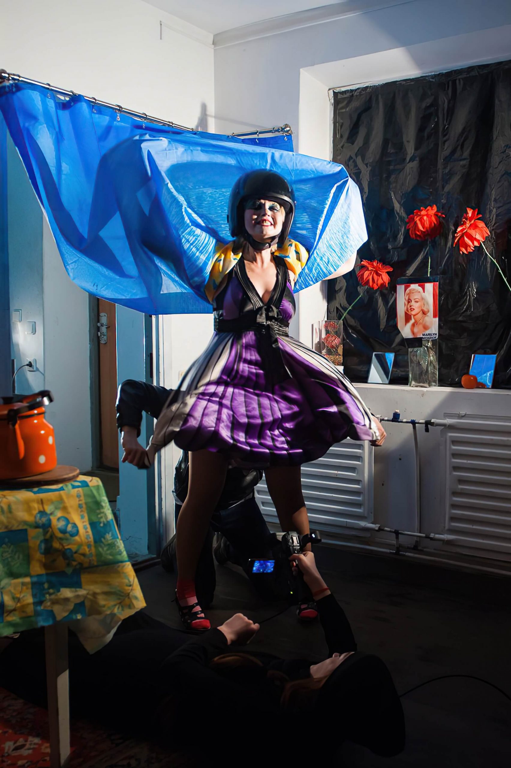 A woman with heavy makeup is wearing a helmet and wrapping herself with a blue curtain