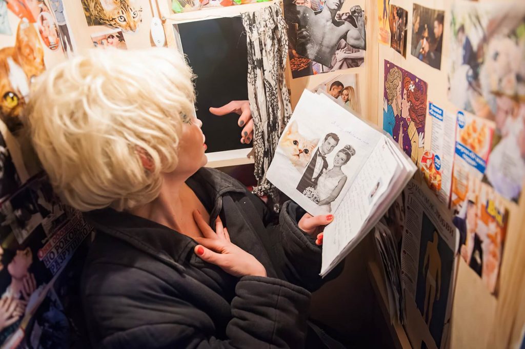 A woman with a heavy makeup reading a book while a hand reach the woman through a small window