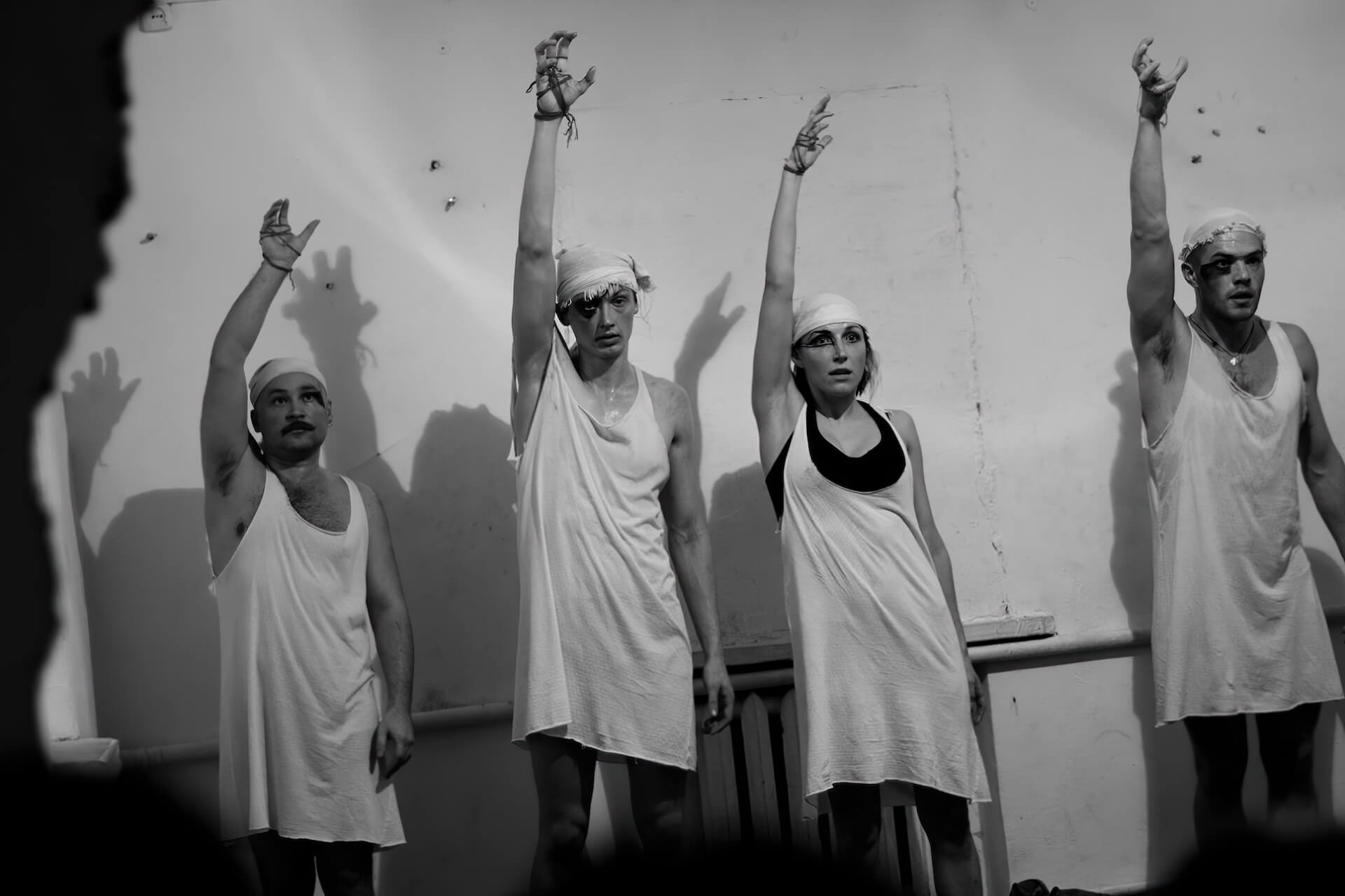 Black and white image of four performers in white garment standing with their grasping hand in the air. Their hands are wrapped around with string and they have unique dark makeup on their right eye.