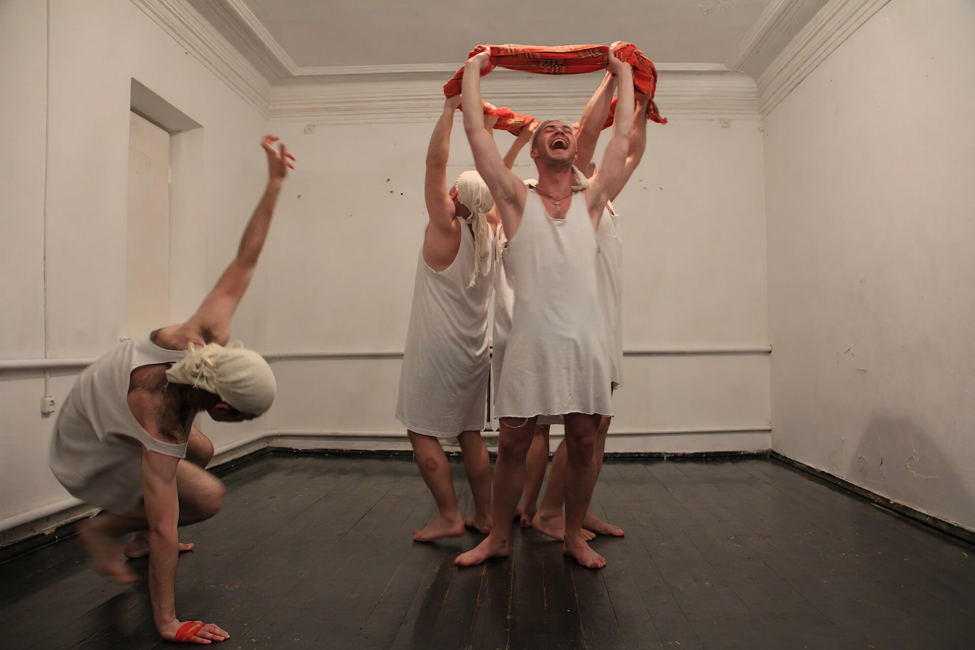 Four performers holding the red blanket up and shouting. One other performer in on the ground with one arm on the floor and the other in the air