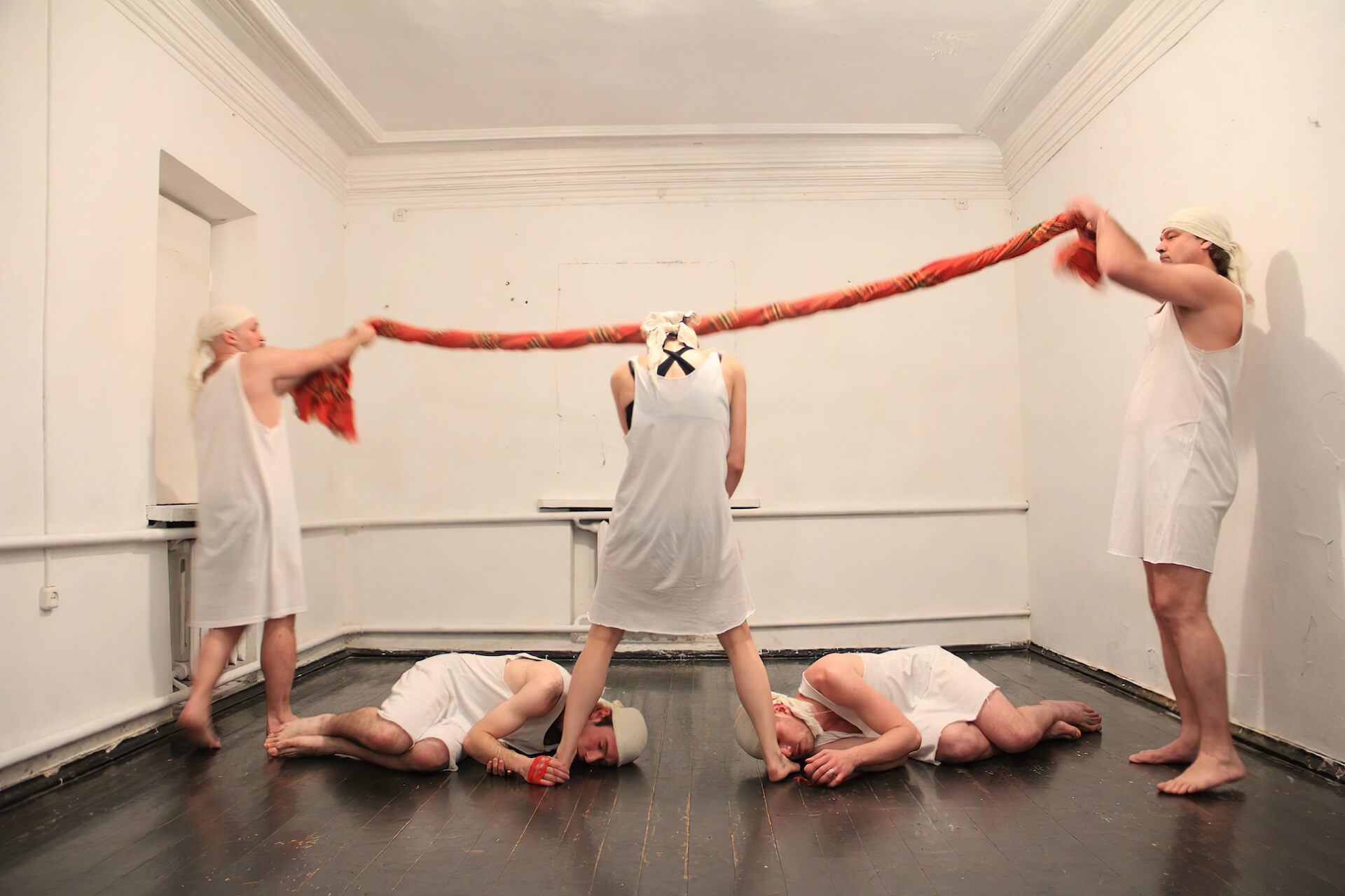 Two performers are standing holding each end of a blanket wrapped around like a rope. In the middle stands a performer and two other performers lying on the floor holding each foot of the performer standing in the middle.