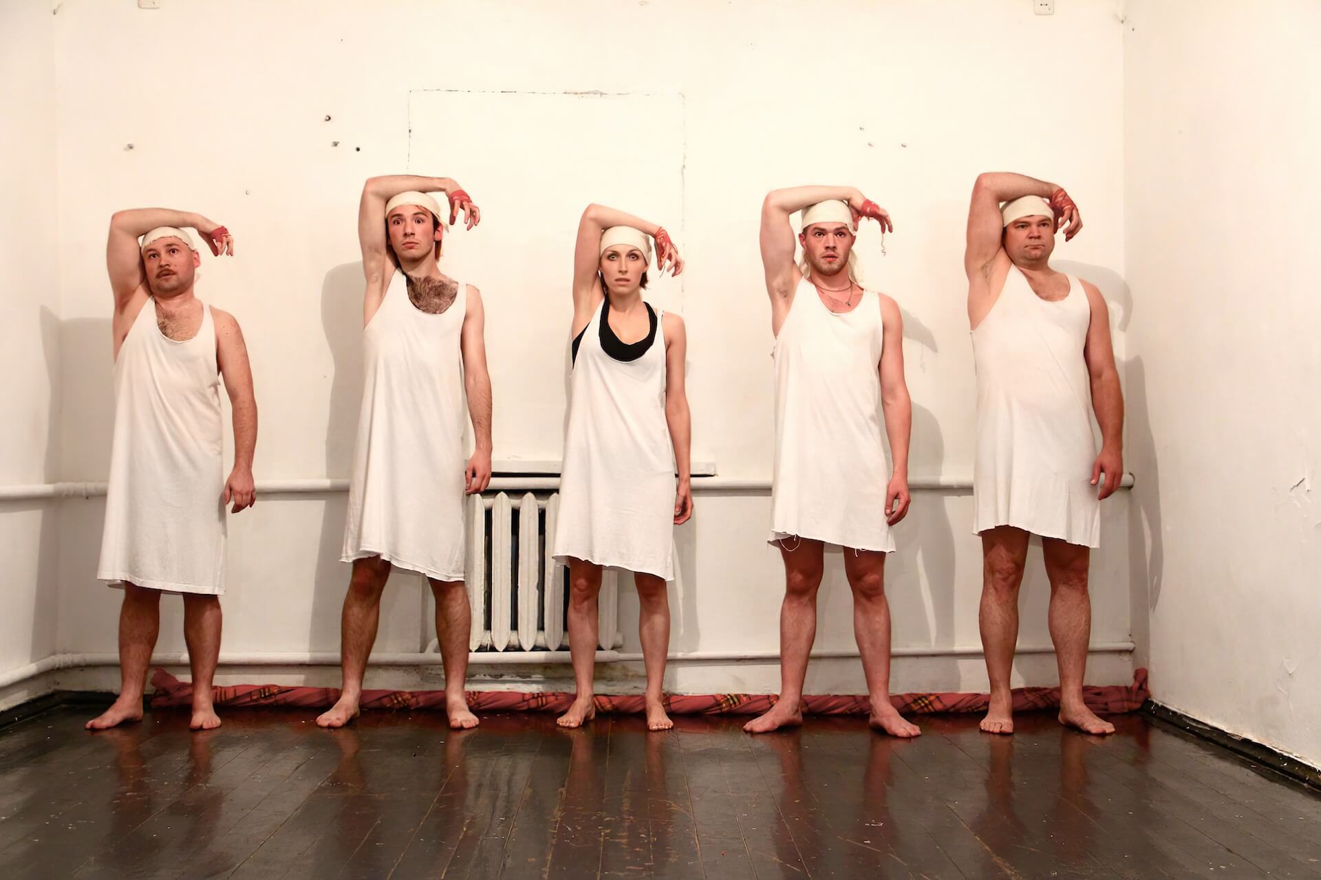 Five performers in white garments are standing in one line with their eyes wide open. They have their right arm on their head.