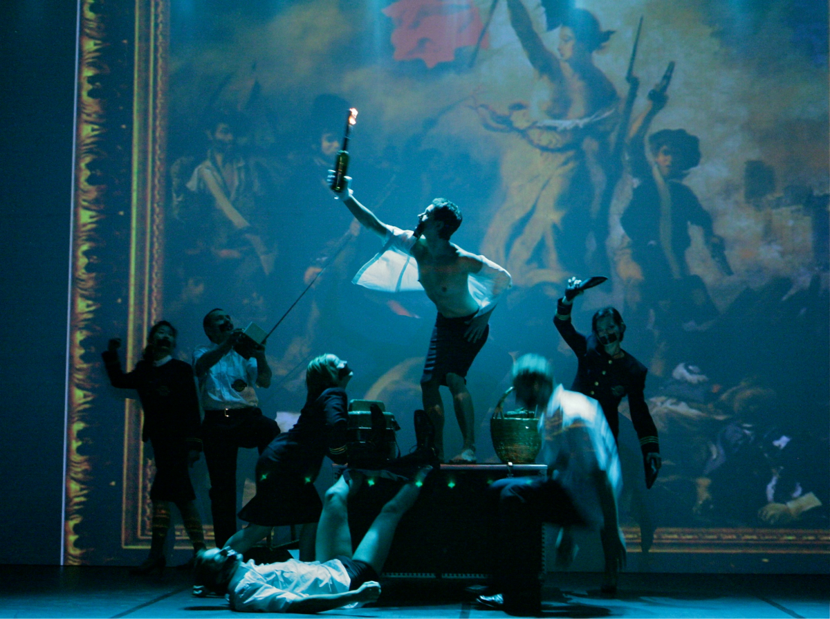 Seven performers are on the stage with their mouths tapes on. In the middle lies a table and one of the performers is standing on it holding up a fire bottle bomb. In the background, there is a huge painting of French revolution