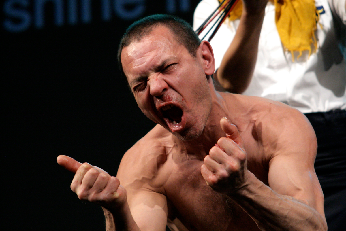 A shirtless performer is grunting. It seems that he has crushed something with both his fists and mouth.