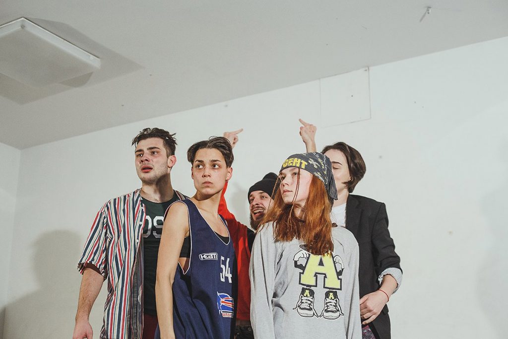Low angle mid shot of five people looking at the front. They are all wearing streetwear and one at the back is holding up both of the middle fingers.