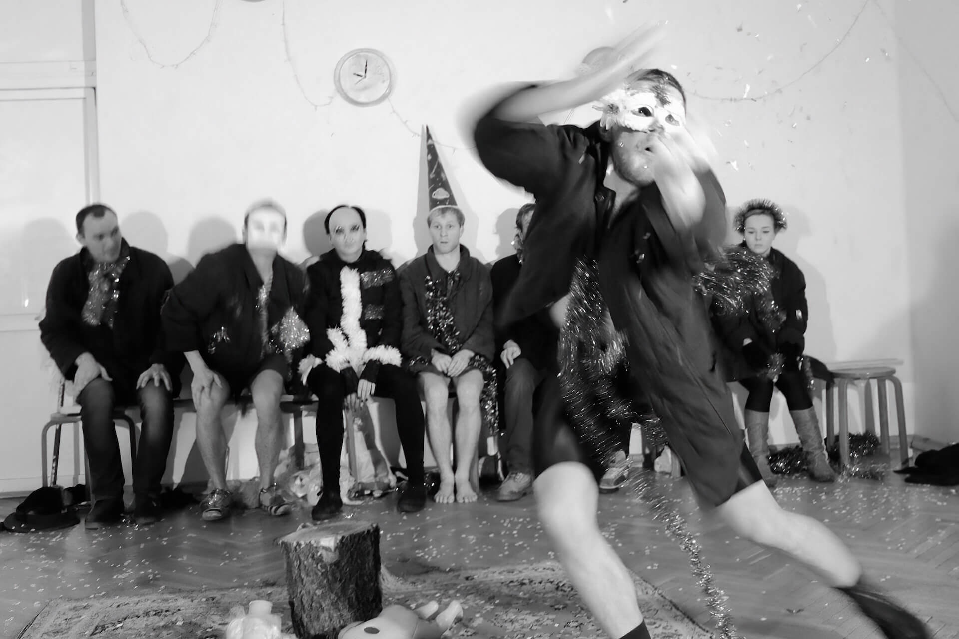 Black and white image of a performer in the middle ripping something sparkly apart vigorously. The others are sitting on a chair by the wall and are watching.