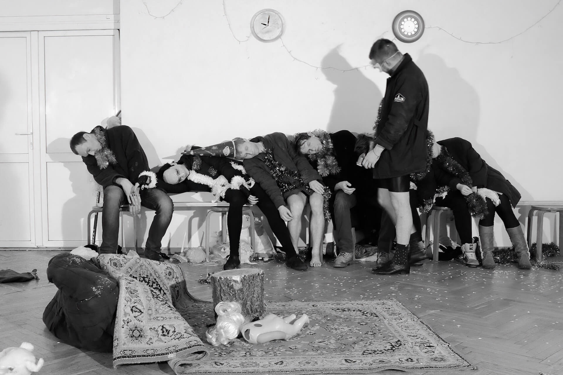 Black and white image of the performers on a chair long the wall sleeping. One other performer is walking around and looking at them.