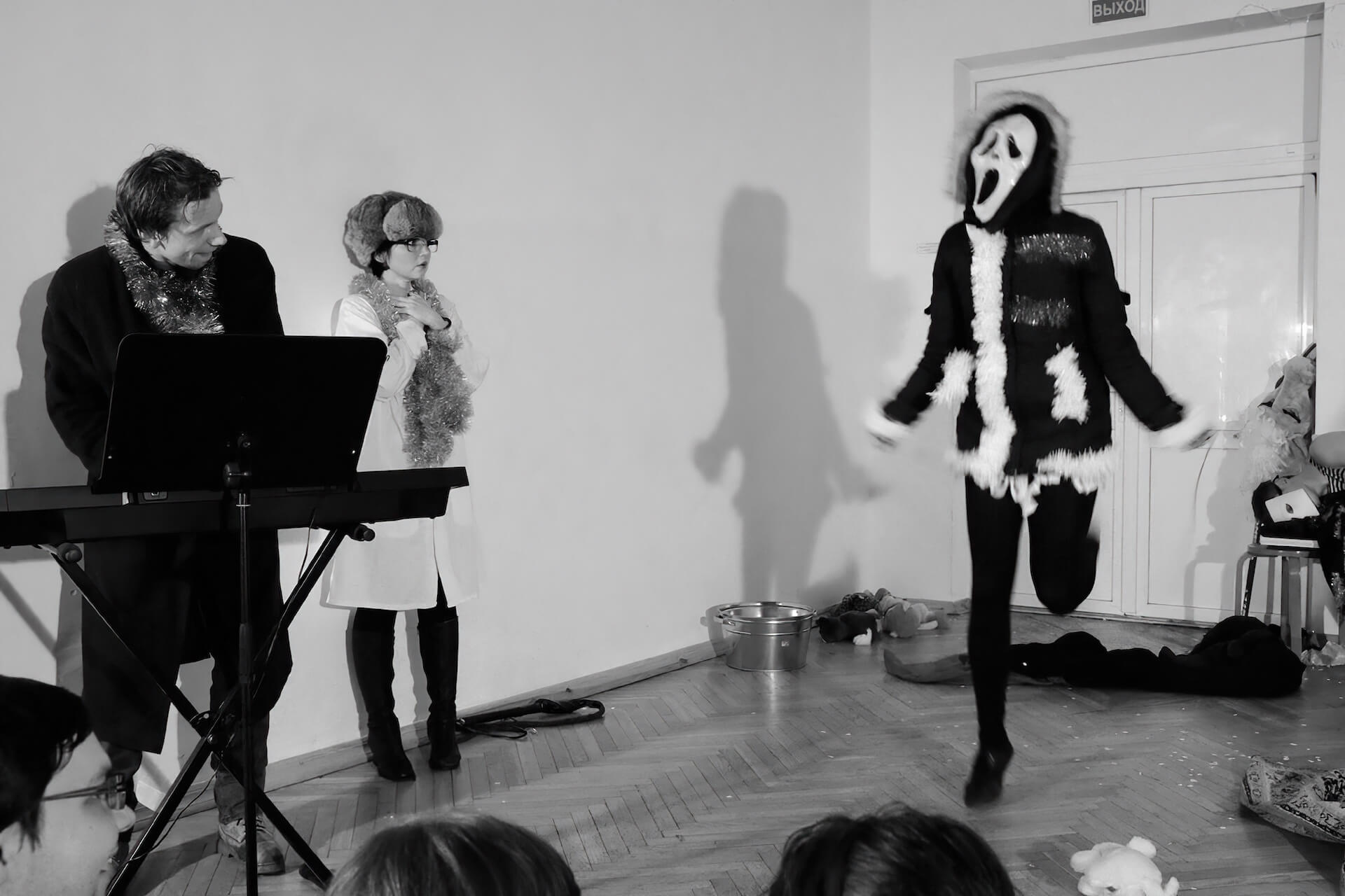 Black and white image of a performer in screaming mask with a father christmas clothing jumping. There is another performer on the side playing keyboard to it.