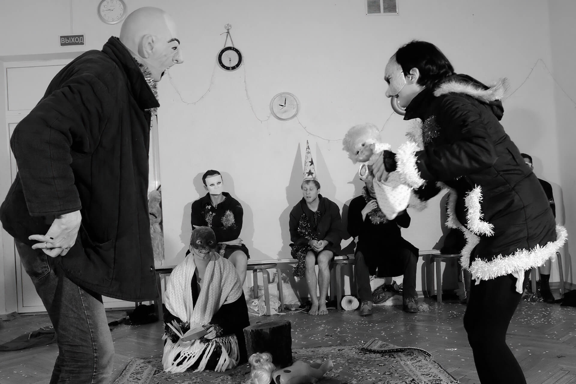 Black and white image of two performers with masks arguing. The rests are sitting behind watching.
