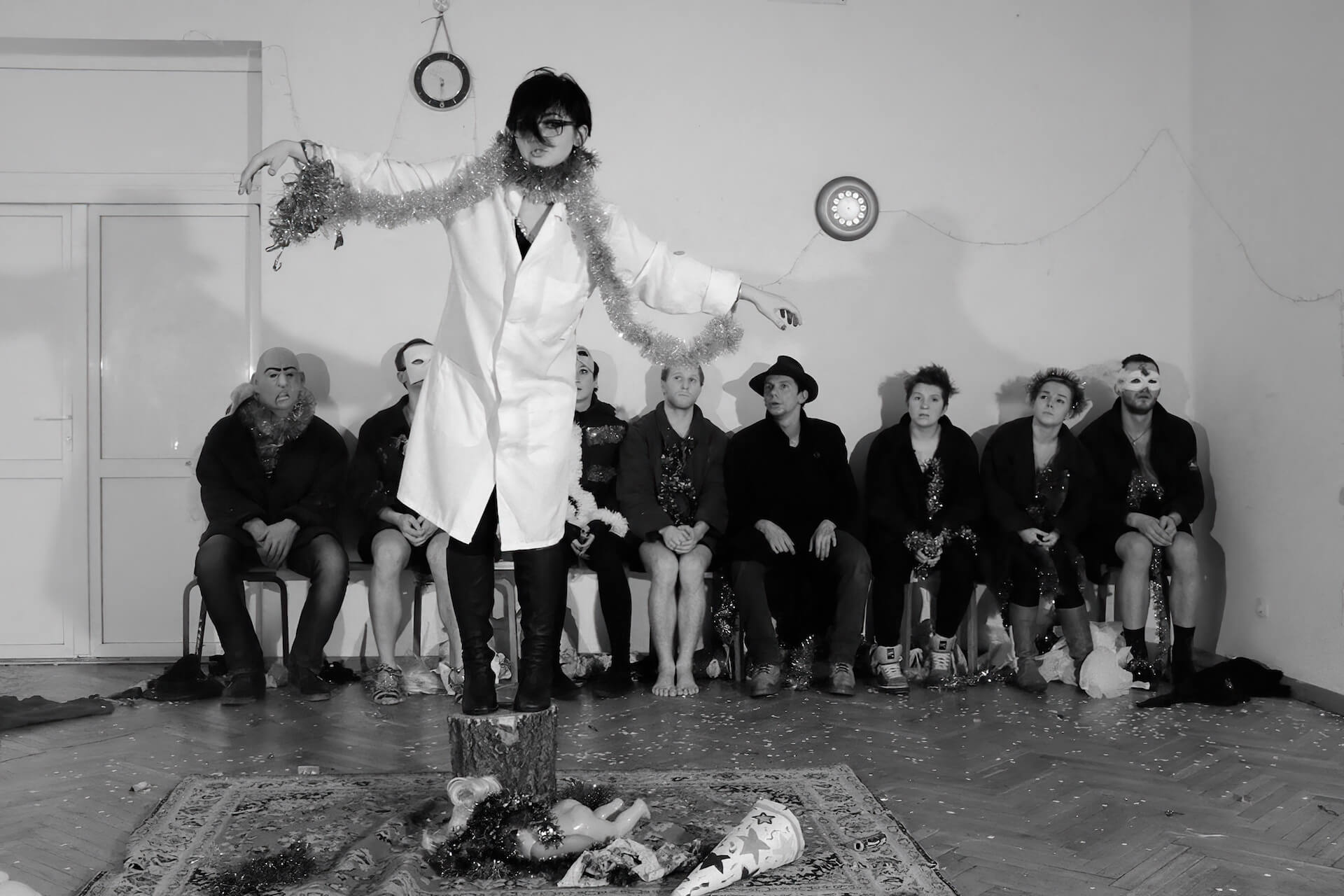 Black and white image of a performer standing on a log in the middle. This performer has handcuffs on the wrists which are linked together by long tinsel. The rest of the performers are sitting in the background on a chair, watching.