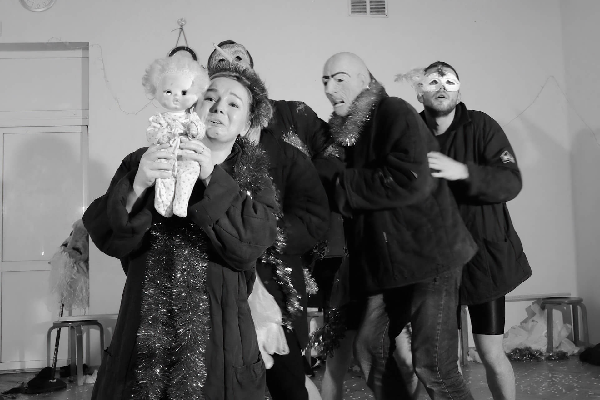 Black and white image of four performers frightened by something and backing up. One other performer is holding up a doll to it with a desperate expression.