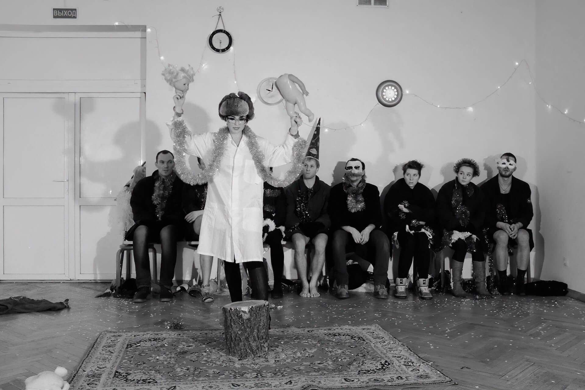 Black and white image of a performer in the middle wearing a ushanka hat. She has handcuffs on her wrists which are linked together with a long tinsel. The performer is holding a broken doll, the head on the right and and the body on the left. Other performers are sitting on a chair looking at the performer in the middle.