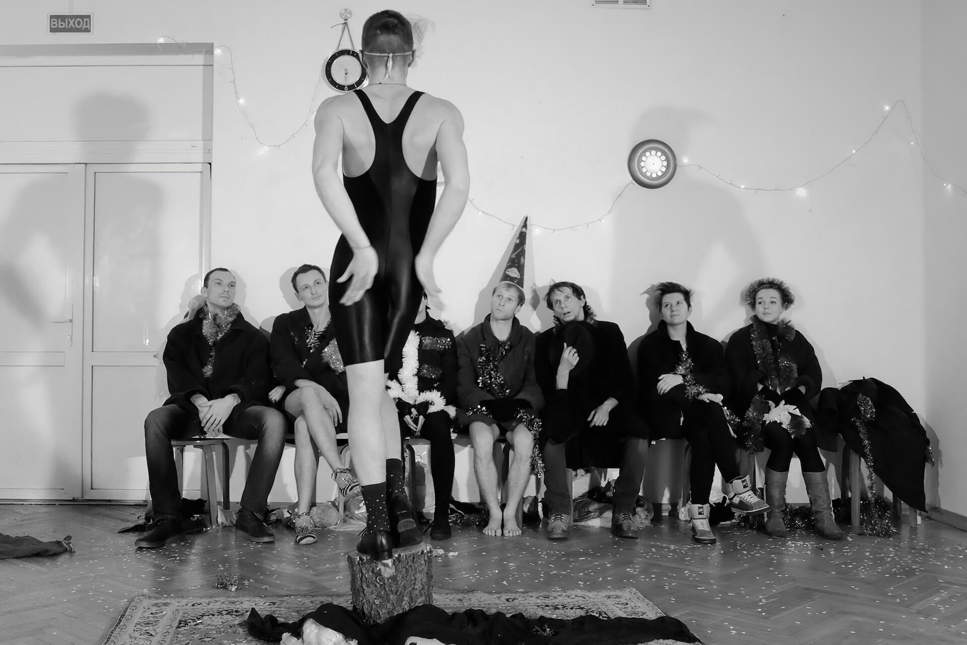 Black and white image of a performer standing in a middle showing off a dance move while the others sit on a chair and watch