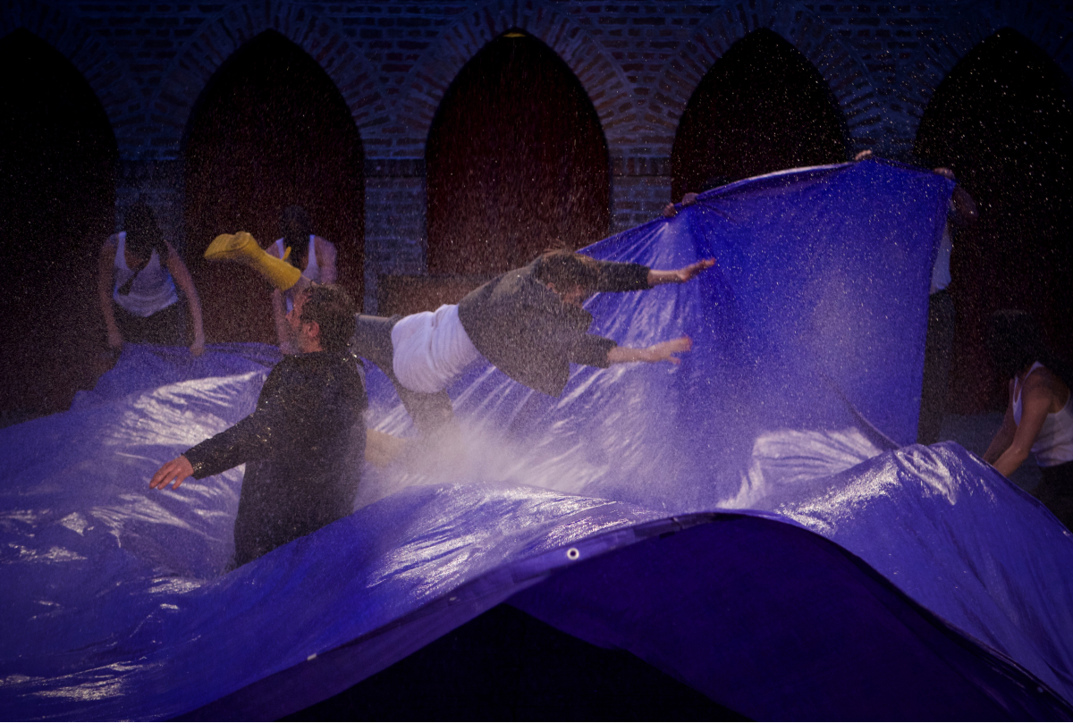 One performer with a pair of yellow rainboots is jumping into the large sheet of blue plastic as if diving into the ocean. Water is splattering from the plastic. In the middle stands a performer looking at the abyss