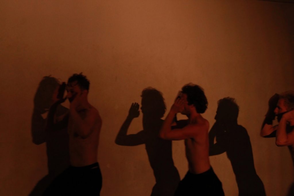 Three performers are running around the dark empty room. They have their hands on each side of their face.