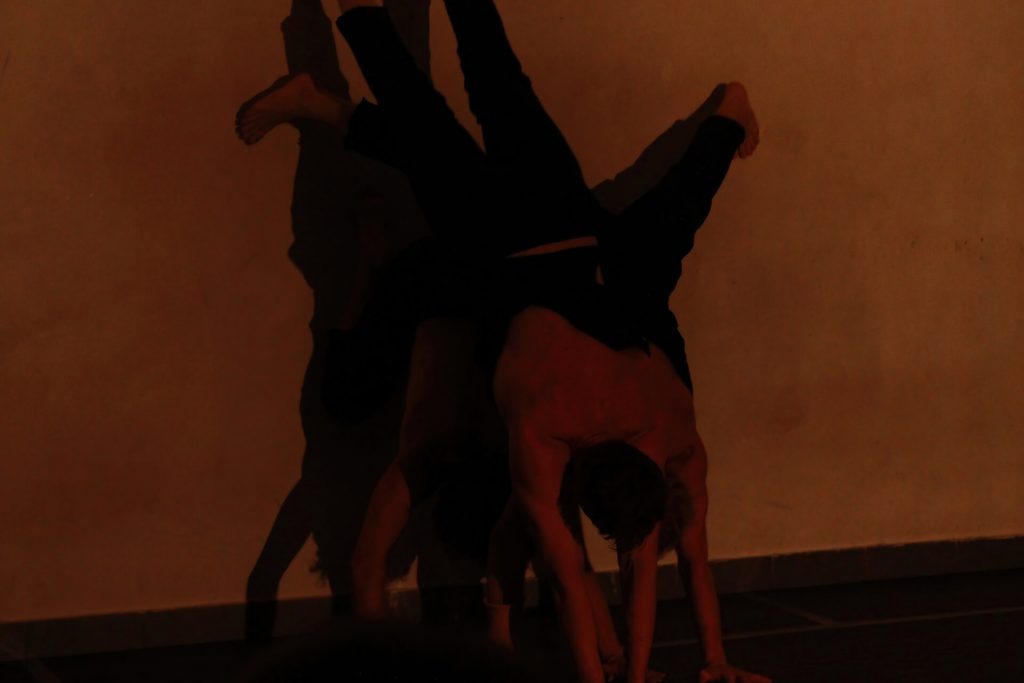 Three performers are in a dark empty room. They are on top of each other, doing handstand against the wall