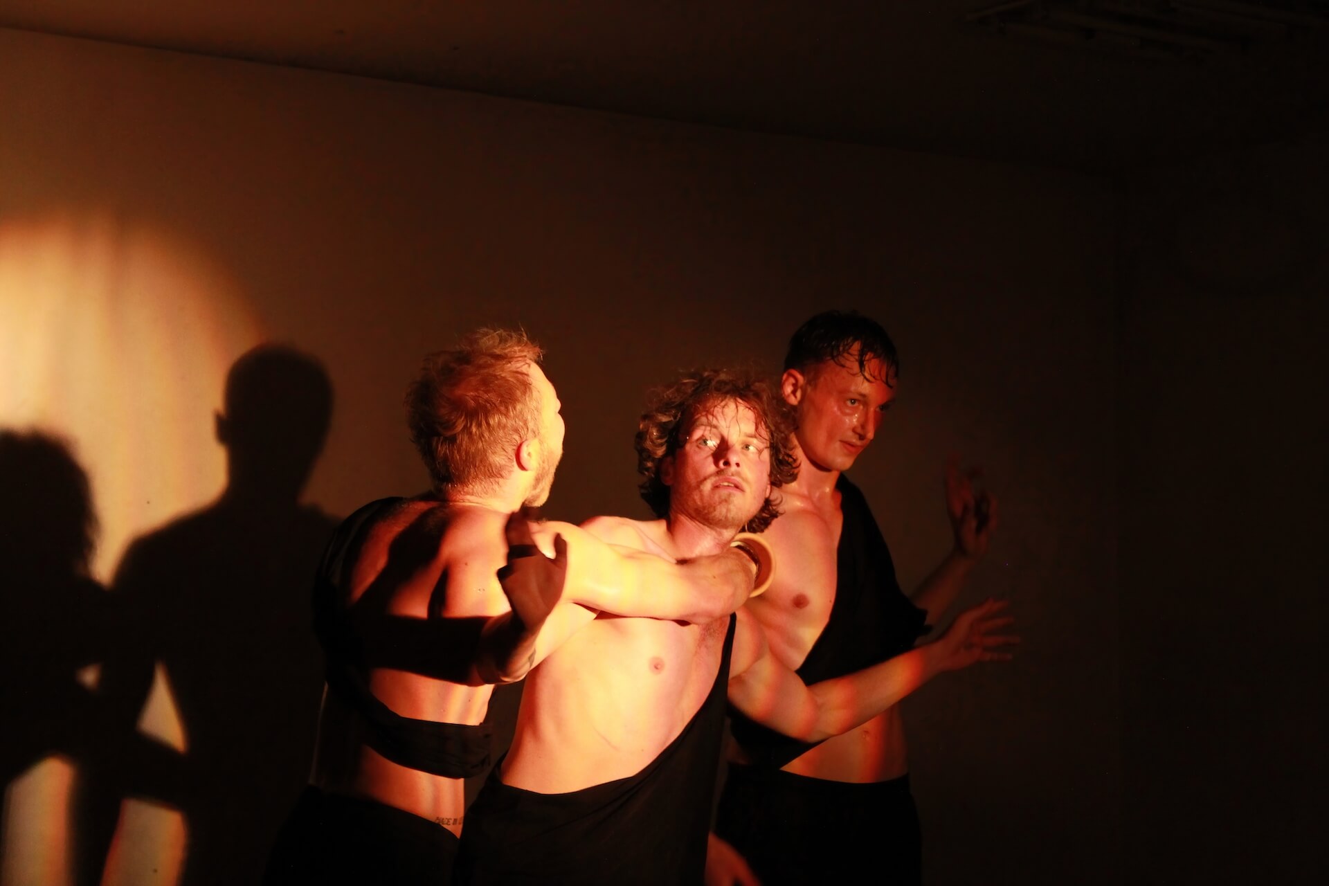 Three performers in a dark empty room. A yellow hinted spotlight shines on them. The one is the middle is looking sideways. The one to his right has his right arm with a vase wrapped around the one in the middle, shouting.