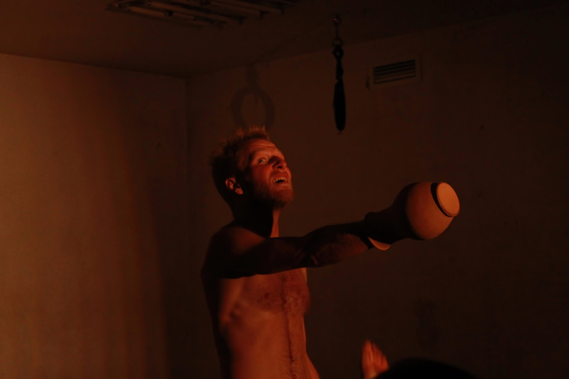 A performer standing in the middle of a dark empty room. He takes his hand stuck in a vase to the front.