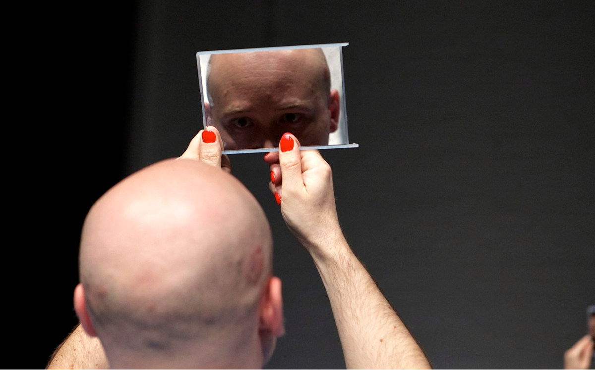 Close up to the back of a bald performer. They have their nails painted in red and are holding up a mirror and looking at their face.