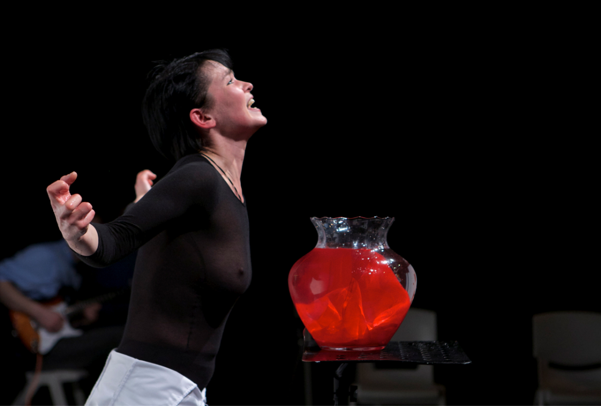 One performer with black sheer tight shirt is shouting at something upwards, with their arms spread out and hands curled up. In front of them, there lies a large glass vase with something red inside.