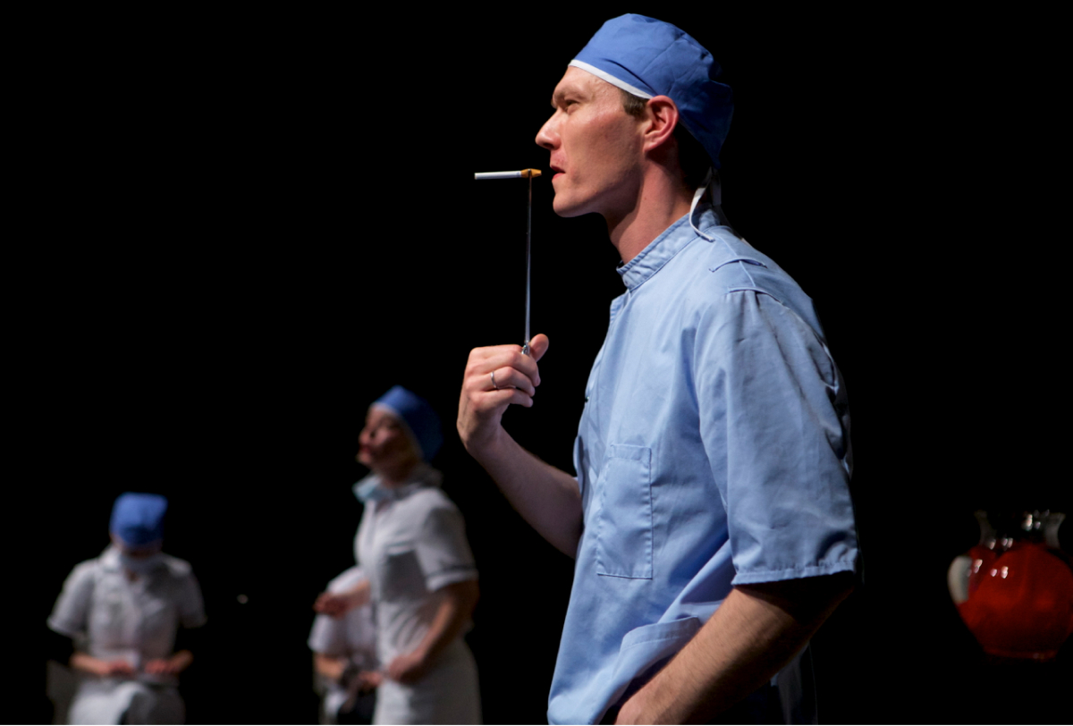 One performer with blue shirt and a blue cap is standing sideways. They look like a surgeon. They are smoking, holding up a cigarette with a pair of surgical tongs.. There are several nurses in the background