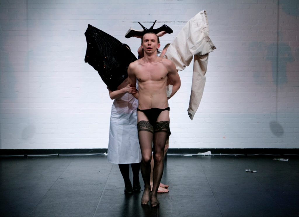 One performer is standing in the middle with their hands tied at the back. They are only wearing underpants and pantyhose. The have a pair of black heels on their head which look like devil horns. They have wings attached at the back made out of clothes. The right wing is formed by a black shiny dress and the left wing is formed by a beige trouser.