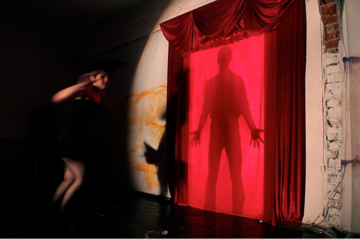 Dark room with a large spotlight on a wall with red stage curtain. In the middle there is a shadow of a person standing. Another performer is running towards that curtain,