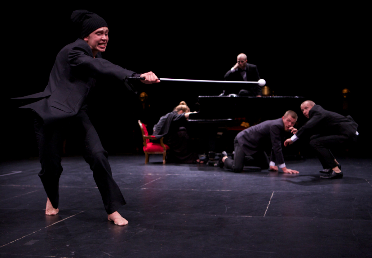 A performer in a black suit is walking barefoot with a black beanie on. With their eyes closed, they are holding a stick with a white golf-sized ball attached at the end. Four people are behind looking at the one in front, looking surprised.