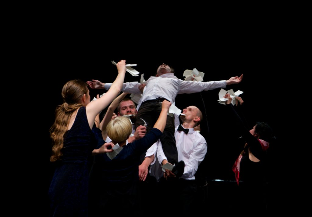 Several performers are in suits and dresses, holding up one performer who has his arms spread out. The ones holding him up is holding pieces of white paper either on their hands or their mouths.