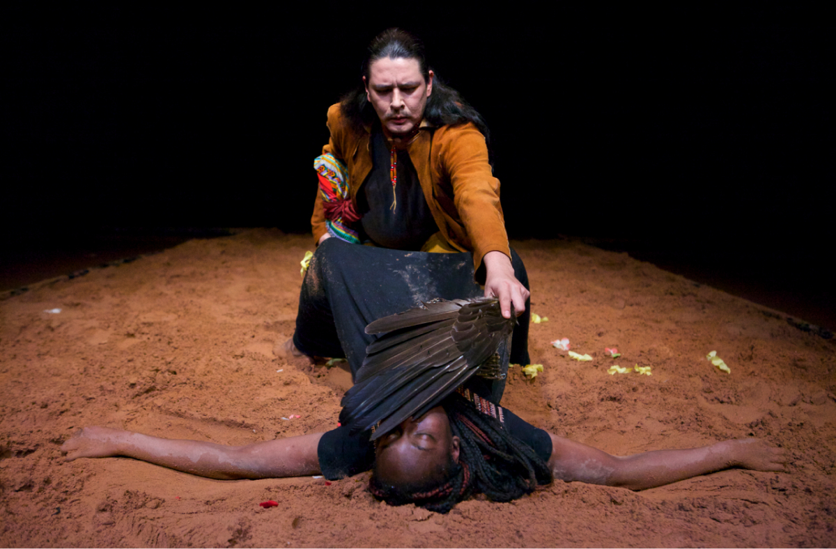 One performer in a black dress is lying on the sand, arms spread out and knees bent. Another performer in a yellow jacket sits between her legs and sweeps the face of the performer lying down with a bundle of feather.