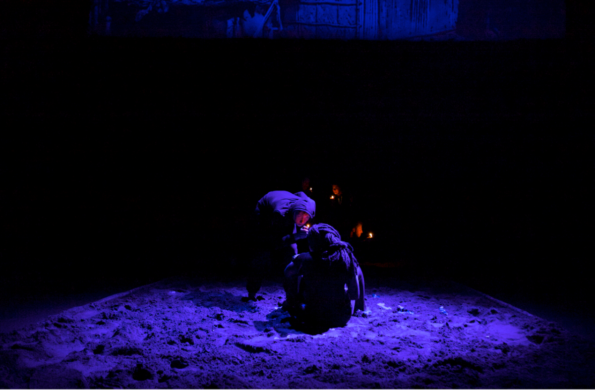 In the darkness with a dim blue light shining on a sand covered floor, a performer with a black dress is sitting down. Another performer stands, bent towards the performer on the ground, holding up a lit up lighter in front of the face of the performer on the ground.