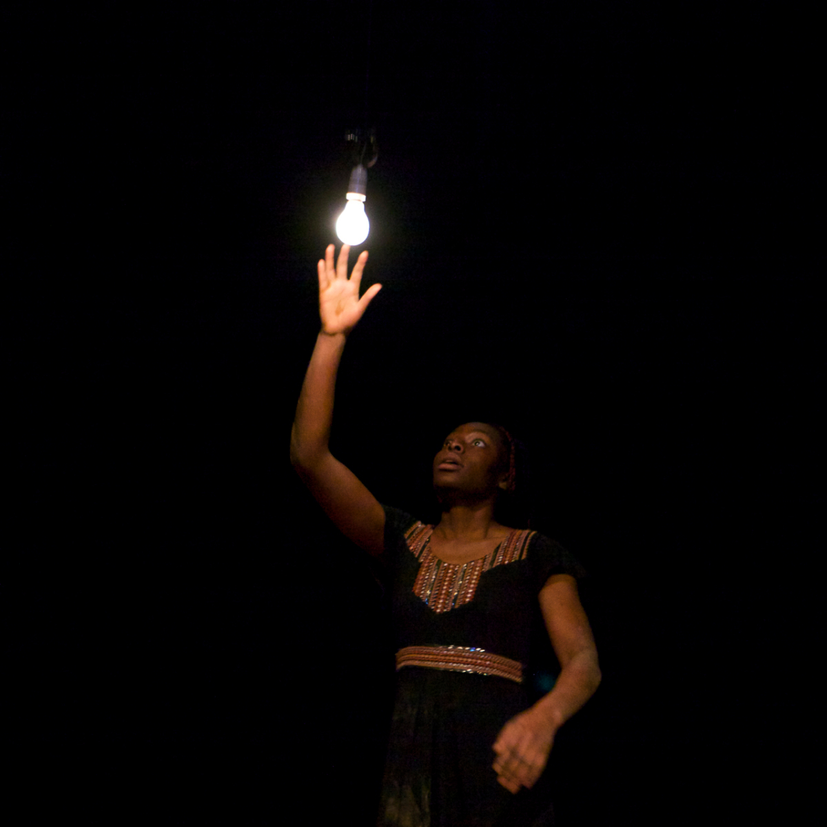 A performer is reaching up for a light bulb hanging on the ceiling. The performer looks surprised or mesmerised.