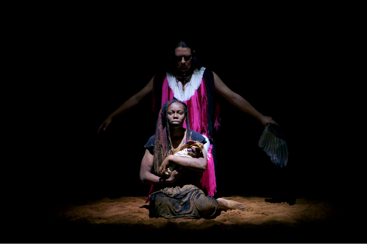 Two performers are on sand. One with a black dress is knelt down holding something in their arms like a baby. Another performer is standing behind with their arms spread out, wearing a dress with pink, red and black fringe. They are holding a bundle of feathers in the left hand.