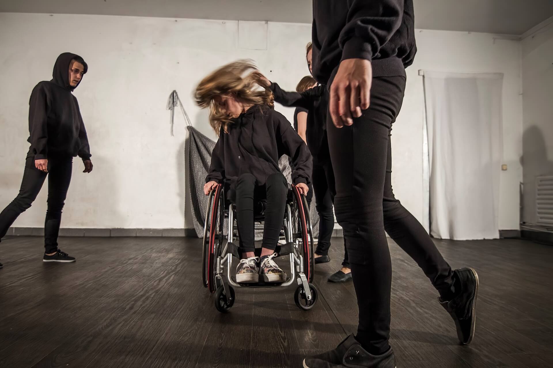 One performer on a wheelchair. Four others are walking around her. The one on the wheelchair is shaking her head vigorously as if she has just been slapped