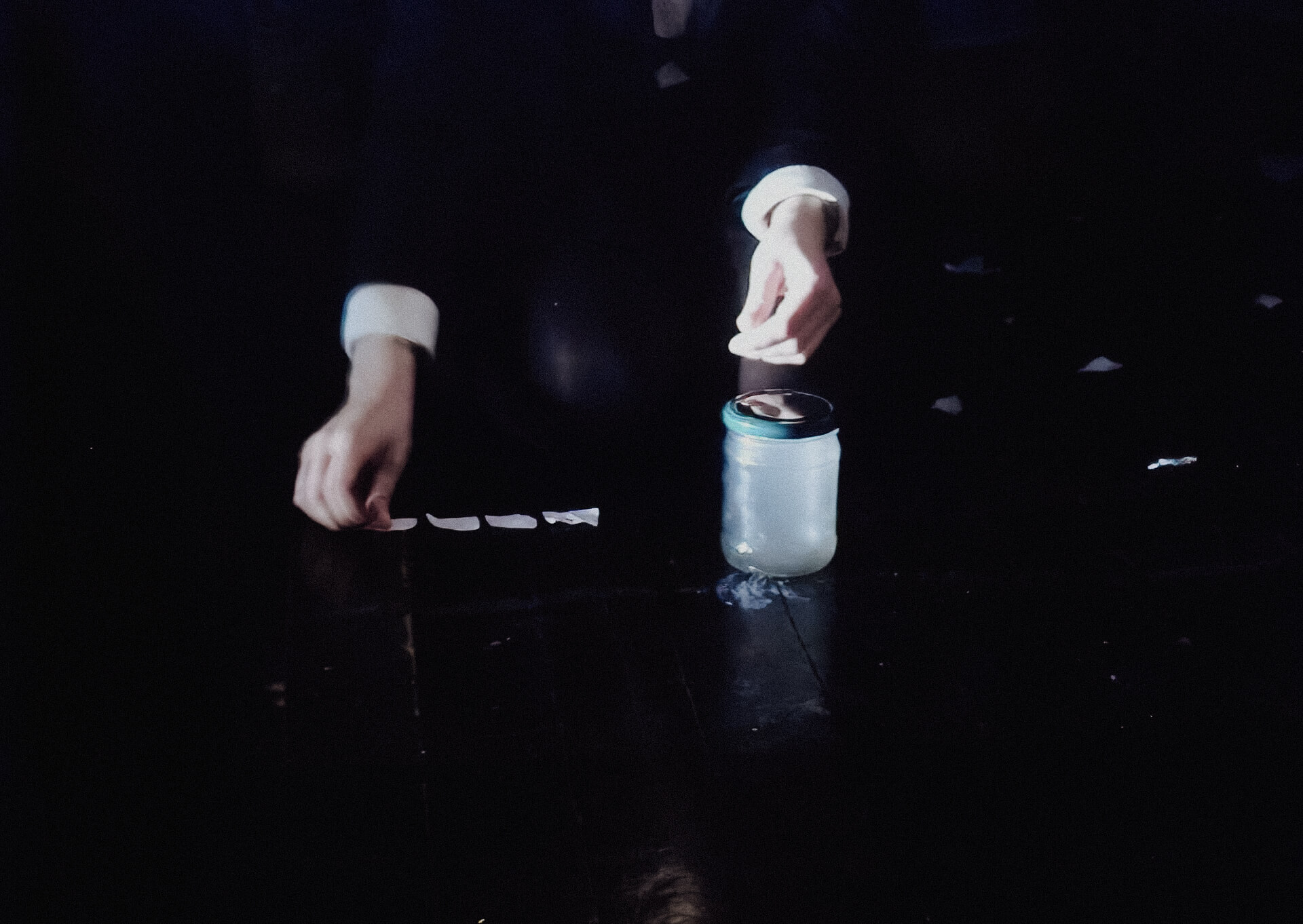 Close up and blurry image of the hands of a performer knelt on the floor. The performer is arranging little piece of paper in one line next to a jar