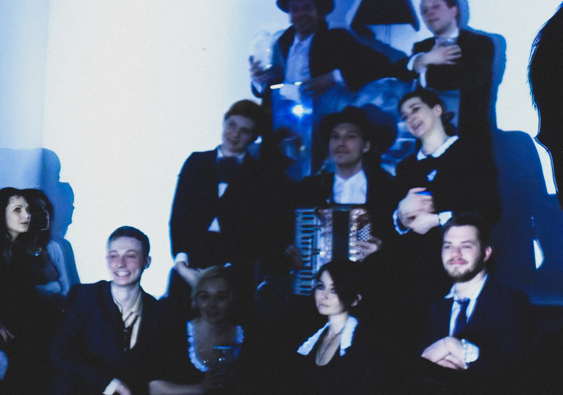 Blurry image of seven performers in black suits and dresses smiling and posing for photo. One is holding an accordion