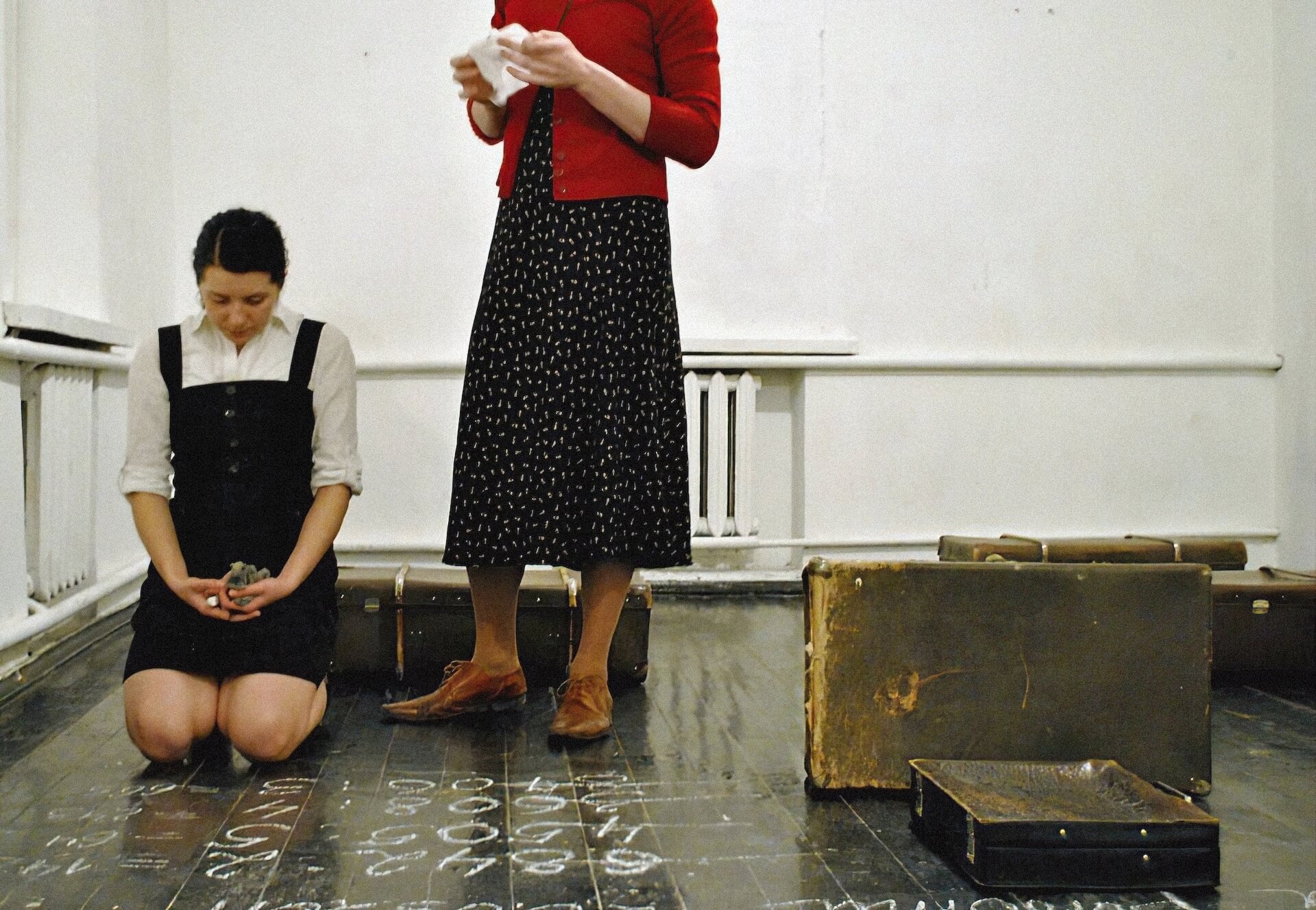A performer in red cardigan, polka dot skirt and yellow knee-high socks is standing next to a knelt down performer. The one on their knee has their head bent forward as if being punished and holding a piece of chalk and cloth. In front of her, there's line of numbers scribbled with chalk.