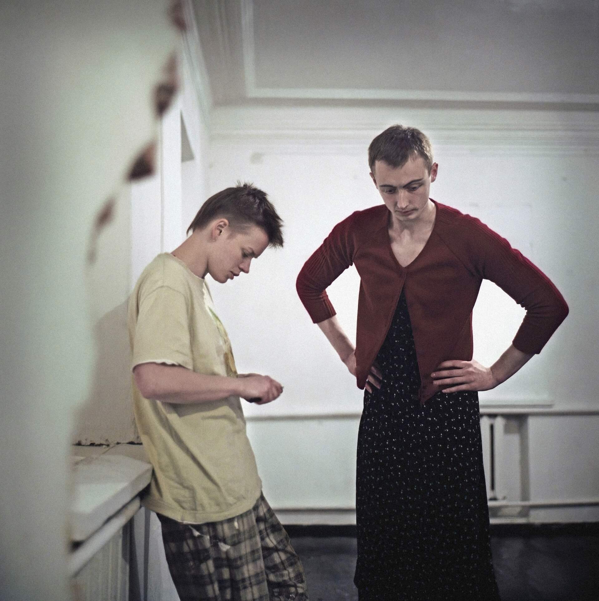 Two people are standing. One in the right is wearing a red cardigan and a polka dot skirt with their arms on the waist. The one on the left is leaning against the wall, looking down.