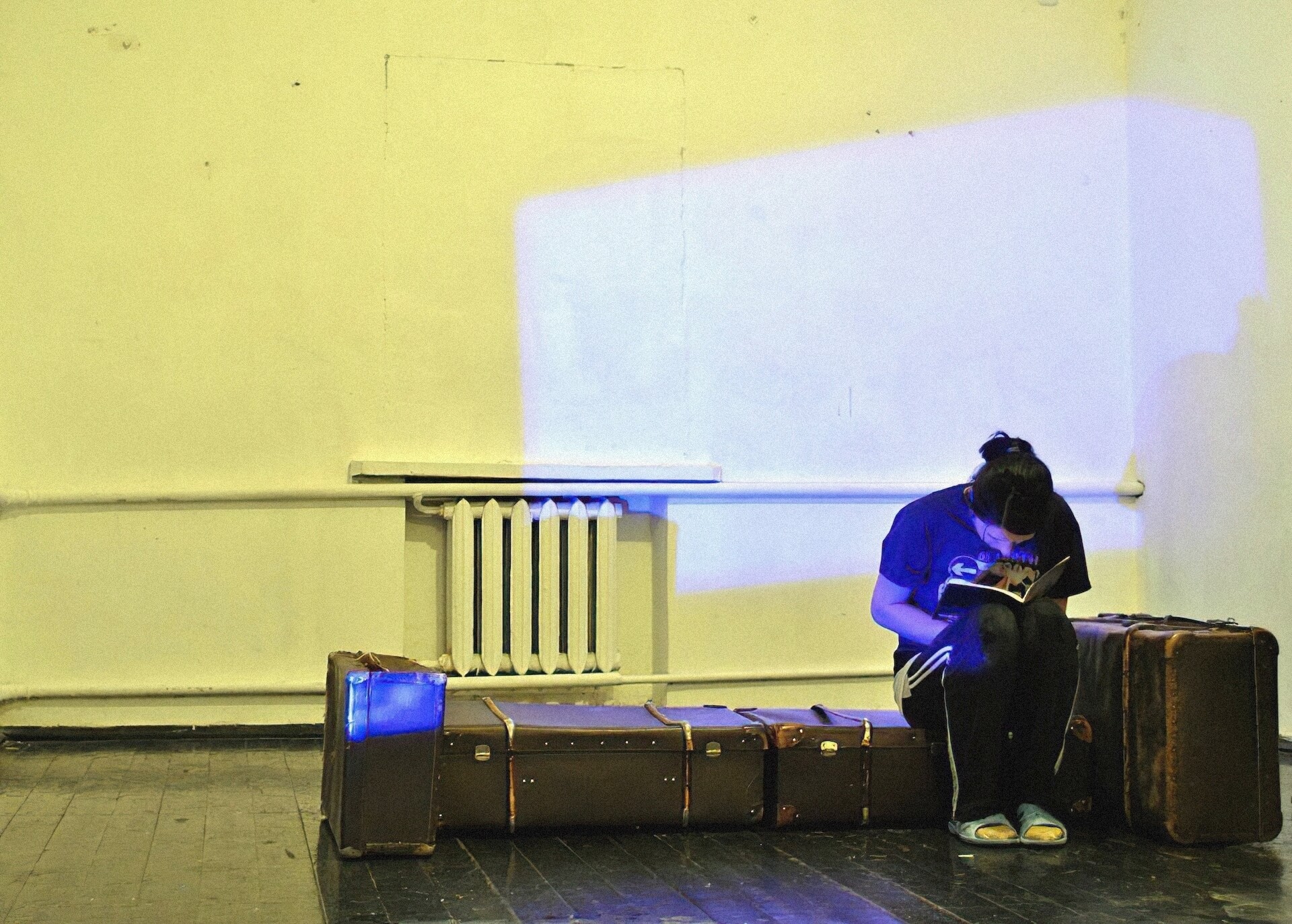 A line of luggage is sitting in an otherwise empty room. Dim blue light is shining on the wall like it's coming from a window. One performer is sitting on a luggage and is reading a book.