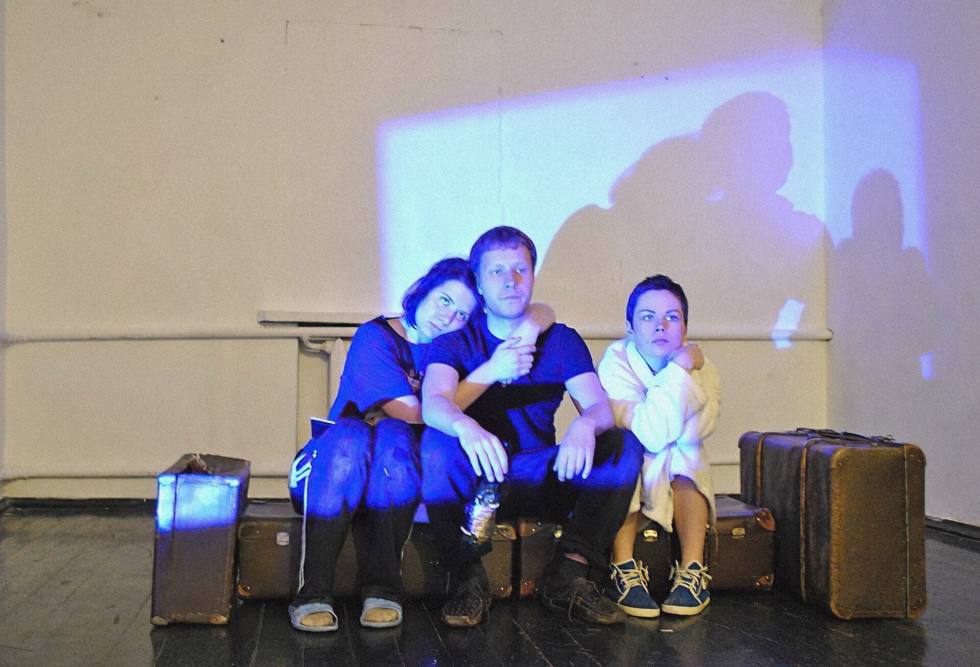 A line of luggage is sitting in an otherwise empty room. Dim blue light is shining on the wall like it's coming from a window. Three people are sitting on the luggage, on on the left wrapping their arms around the one in the middle. All three of them are looking at something in front, unamused