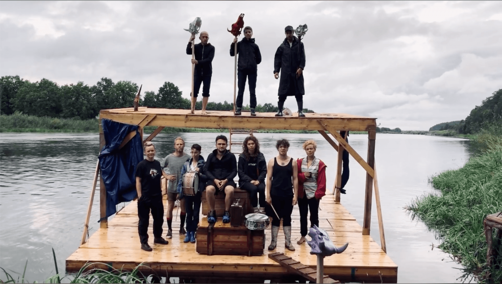 A wooden raft is floating on a river. Ten performers are standing on the raft looking at the camera