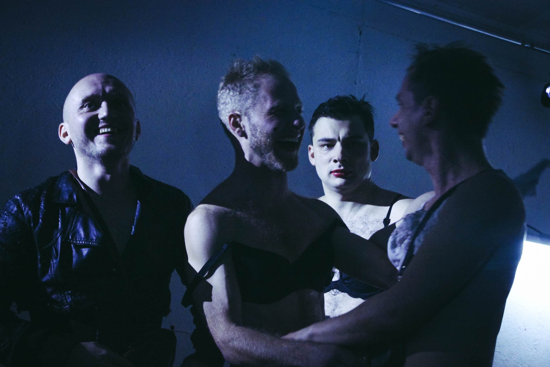 In a dark blue lighted room, four performers are smiling and hugging each other except for one.