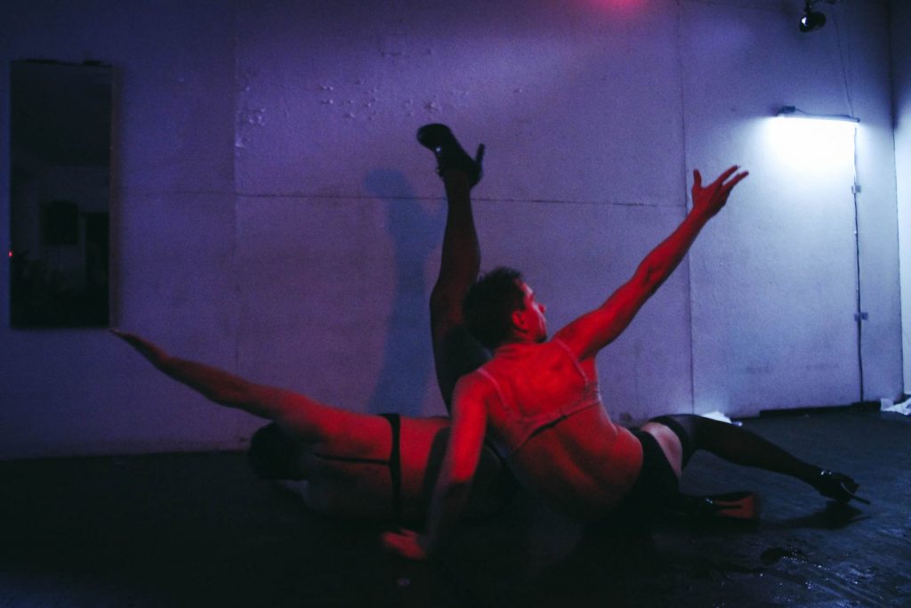 In a dark blue lighted room two performers are shined with red light. They are wearing a bra, underpants, pantyhose and high heels. One is lying down with the right arm and right leg in the air while the other is sitting sideways with the right arm reaching upwards.