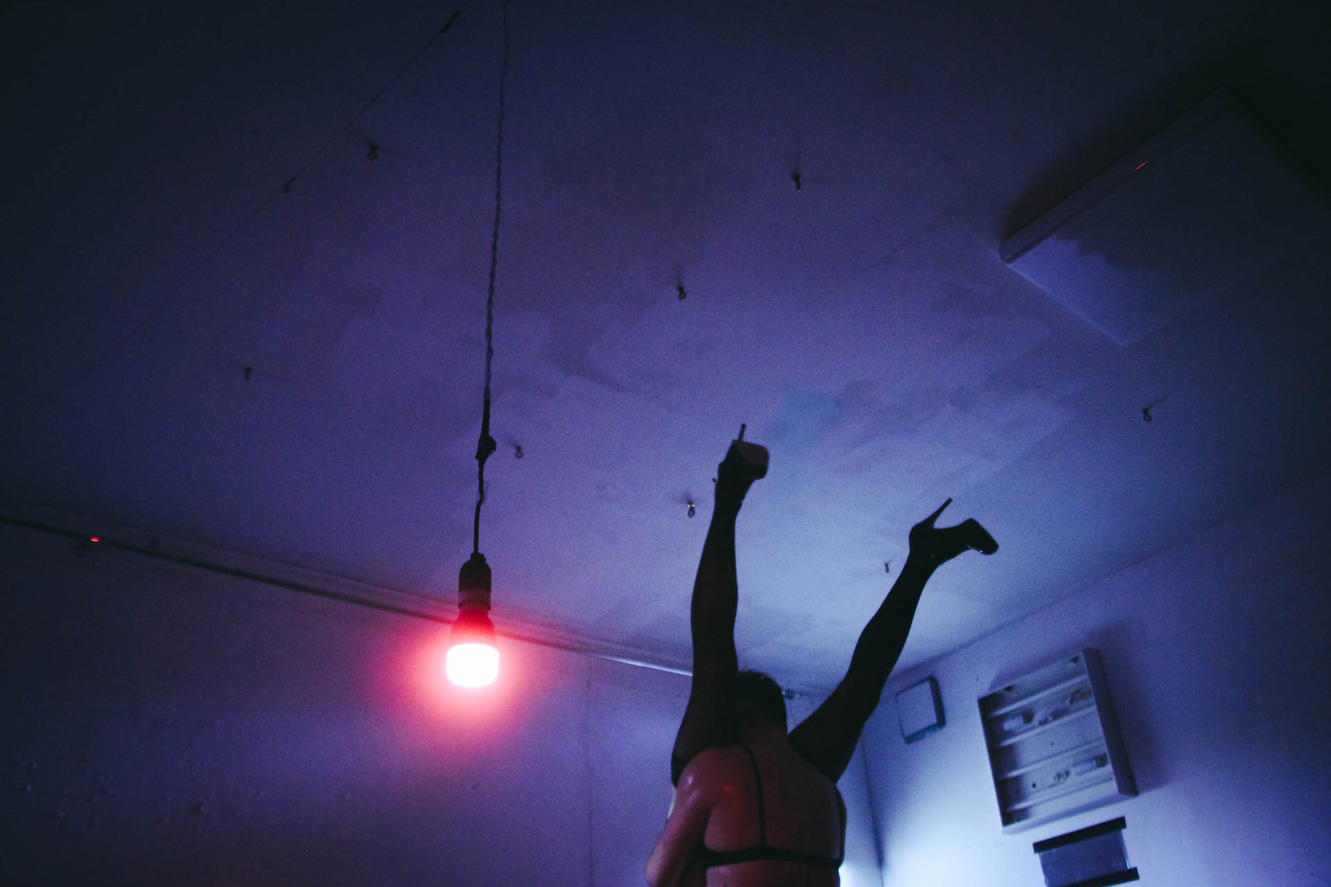In a dark blue lighted room, a red light bulb is hanging by the ceiling. One performer wearing a bra is holding another performer wearing pantyhose and high heels up-side-down by the waist.