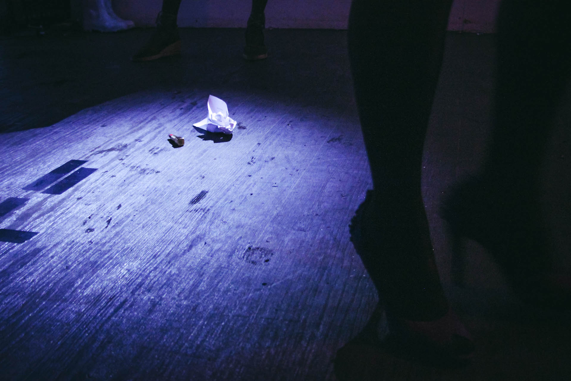 Zoom in to the floor in a dark blue lighted room. There is a silhouette of a performer's legs with high heels. On the floor there lies a lipstick and a crumpled piece of paper.